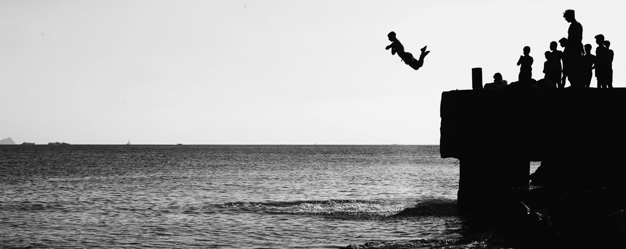 Group of people silhouetted against the sea, with one jumping from a pier in a spontaneous act of fun.