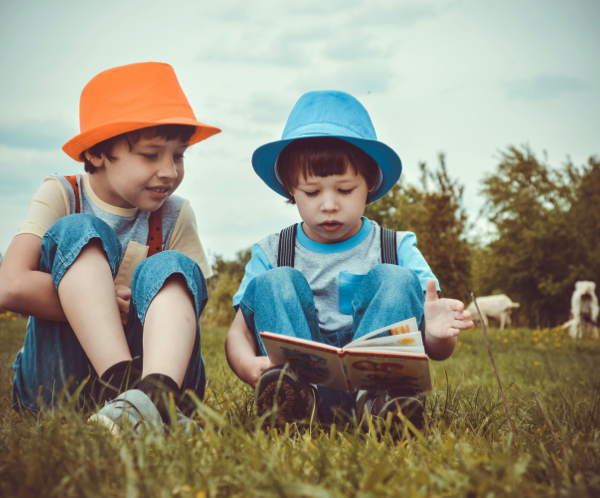 Two young boys enjoying a book together outdoors on a sunny day, wearing vibrant hats.