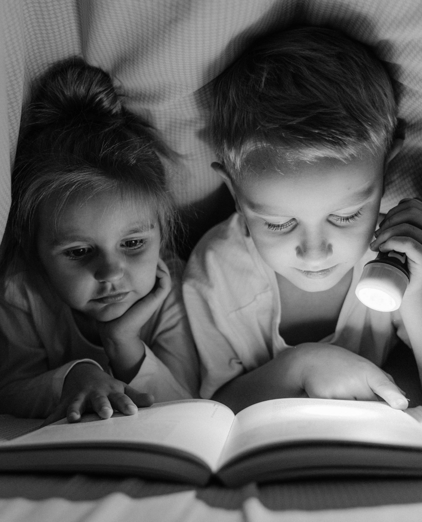 A boy and girl reading with a flashlight under a blanket in grayscale